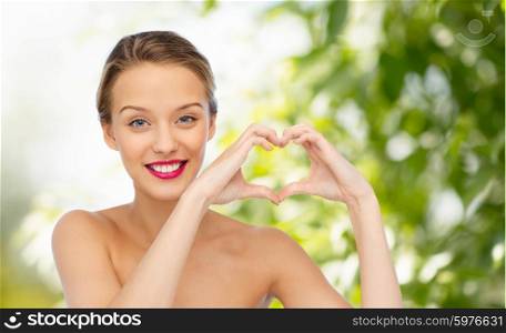 beauty, people, love, valentines day and make up concept - smiling young woman with pink lipstick on lips showing heart shape hand sign over green natural background
