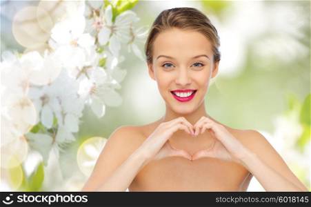 beauty, people, love, valentines day and make up concept - smiling young woman with pink lipstick on lips showing heart shape hand sign over green natural background with cherry blossom