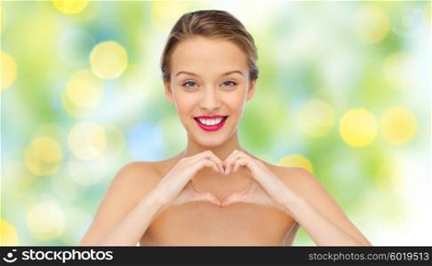 beauty, people, love, valentines day and make up concept - smiling young woman with pink lipstick on lips showing heart shape hand sign over green lights background