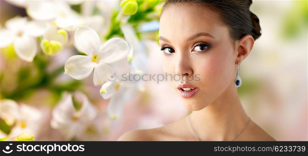 beauty, people, make up and luxury concept - close up of beautiful asian woman or bride with earring over natural spring lilac blossom background