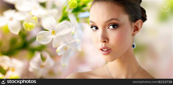 beauty, people, make up and luxury concept - close up of beautiful asian woman or bride with earring over natural spring lilac blossom background