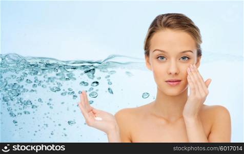 beauty, people, moisturizing cosmetics, skin care and health concept - young woman applying cream to her face over water splash background
