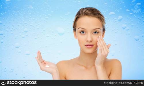 beauty, people, moisturizing cosmetics, skin care and health concept - young woman applying cream to her face over water drops on blue background
