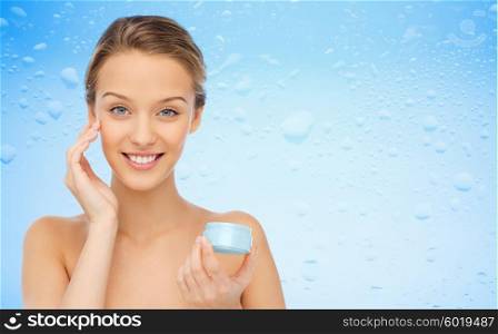 beauty, people, moisturizing, skin care and cosmetics concept - happy young woman applying cream to her face over water drops on blue background
