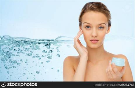 beauty, people, moisturizing, skin care and cosmetics concept - young woman applying cream to her face over water splash background