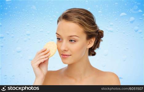 beauty, people, moisturizing, skin care and skincare concept - young woman cleaning face with exfoliating sponge over water drops on blue background