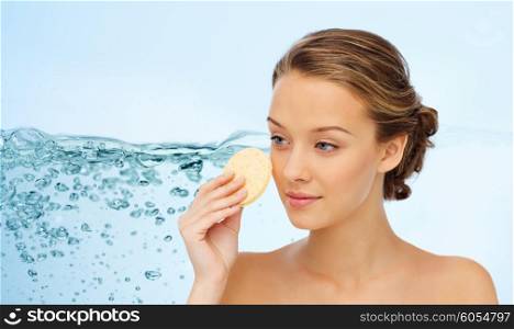 beauty, people, moisturizing, skin care and skincare concept - young woman cleaning face with exfoliating sponge over water splash background