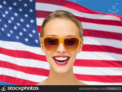 beauty, people, nationality and patriotism concept - smiling young woman in sunglasses with pink lipstick on lips over american flag background