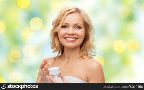 beauty, people, skincare and cosmetics concept - happy middle aged woman with cream jar over green holidays lights background