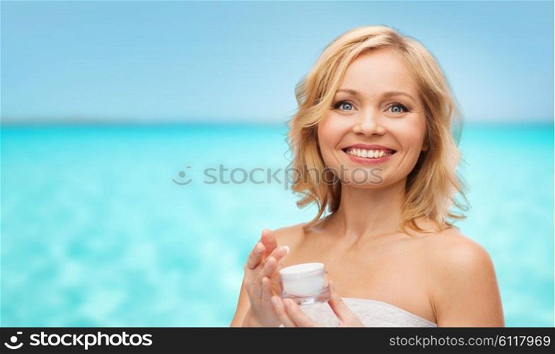 beauty, people, skincare and cosmetics concept - happy middle aged woman with cream jar over blue sea and sky background