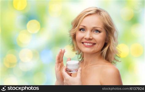 beauty, people, skincare and cosmetics concept - happy woman applying cream to her face over green summer lights background