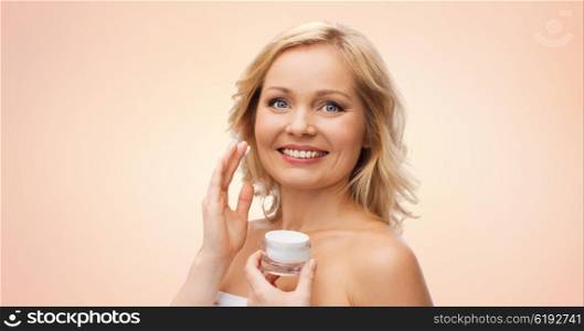 beauty, people, skincare and cosmetics concept - happy woman applying cream to her face over beige background