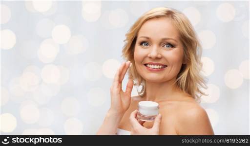 beauty, people, skincare and cosmetics concept - happy woman applying cream to her face over holidays lights background