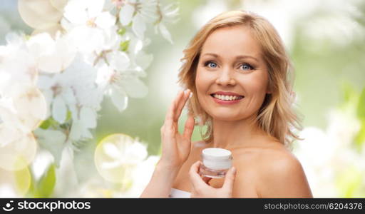 beauty, people, skincare and cosmetics concept - happy woman applying cream to her face over natural spring cherry blossom background