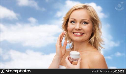 beauty, people, skincare and cosmetics concept - happy woman applying cream to her face over blue sky and clouds background