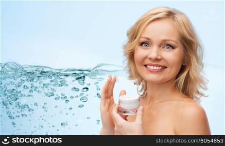 beauty, people, skincare and cosmetics concept - happy woman applying moisturizing cream to her face over blue background with water splash