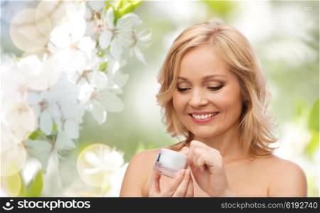 beauty, people, skincare and cosmetics concept - happy woman with cream jar over natural spring cherry blossom background