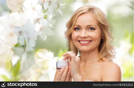 beauty, people, skincare and cosmetics concept - happy woman with cream jar over natural spring cherry blossom background