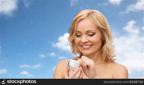 beauty, people, skincare and cosmetics concept - happy woman with cream jar over blue sky and clouds background