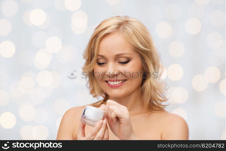 beauty, people, skincare and cosmetics concept - happy woman with cream jar over holidays lights background