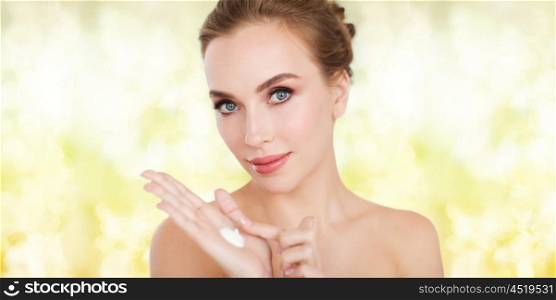 beauty, people, skincare and cosmetics concept - happy young woman with moisturizing cream on hand over yellow holidays lights background