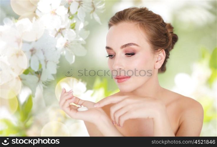 beauty, people, skincare and cosmetics concept - happy young woman with moisturizing cream on hand over natural spring cherry blossom background