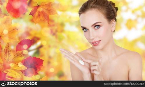 beauty, people, skincare and cosmetics concept - happy young woman with moisturizing cream on hand over natural autumn leaves and lights background