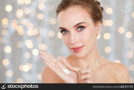 beauty, people, skincare and cosmetics concept - happy young woman with moisturizing cream on hand, over holidays lights background. woman with moisturizing cream on hand over lights