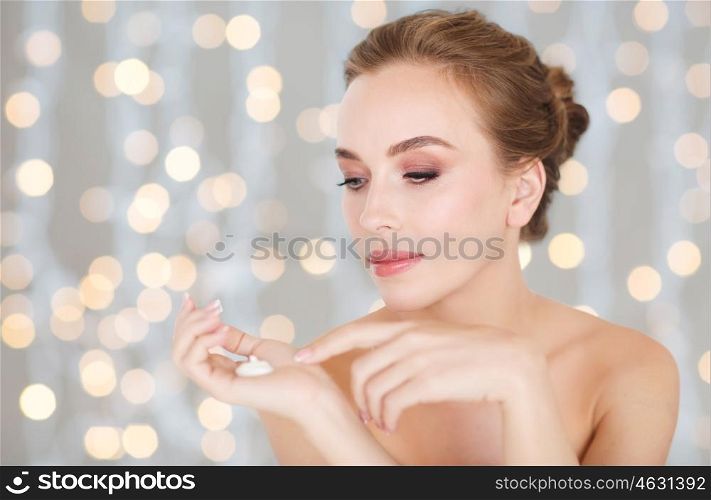 beauty, people, skincare and cosmetics concept - happy young woman with moisturizing cream on hand, over holidays lights background
