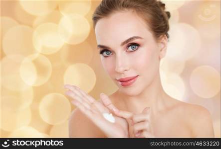 beauty, people, skincare and cosmetics concept - happy young woman with moisturizing cream on hand, over holidays lights background