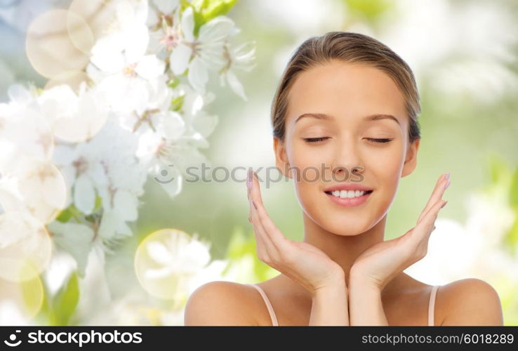 beauty, people, skincare and health concept - smiling young woman face and hands over green natural background with cherry blossom