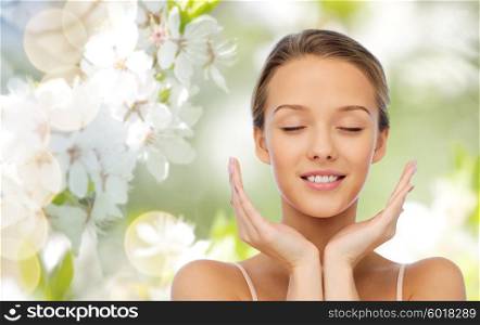 beauty, people, skincare and health concept - smiling young woman face and hands over green natural background with cherry blossom