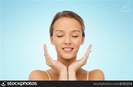 beauty, people, skincare and health concept - smiling young woman face and hands over blue background