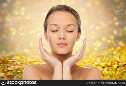 beauty, people, skincare and health concept - young woman face and hands over golden holidays lights or yellow glitter background