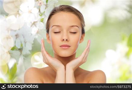 beauty, people, skincare and health concept - young woman face and hands over cherry blossom background