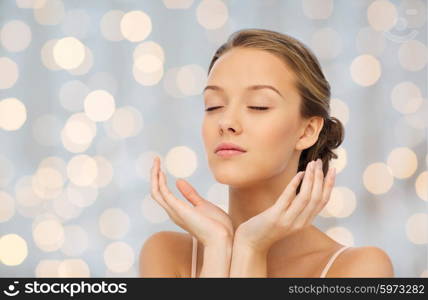 beauty, people, skincare and health concept - young woman face and hands over holidays lights background