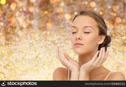 beauty, people, skincare and health concept - young woman face and hands over holidays lights background