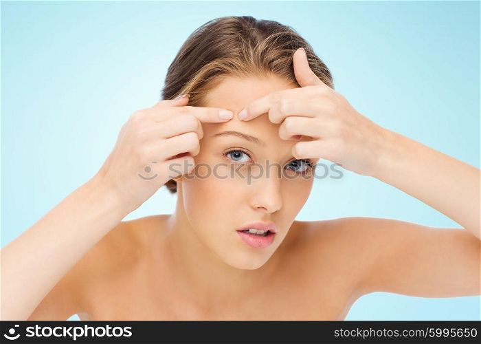 beauty, people, skincare and health concept - young woman squeezing pimple on her face over blue background