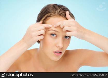 beauty, people, skincare and health concept - young woman squeezing pimple on her face over blue background