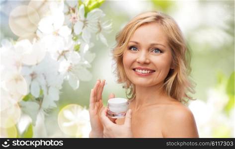 beauty, people, skincare and natural cosmetics concept - happy woman applying cream to her face over cherry blossom background