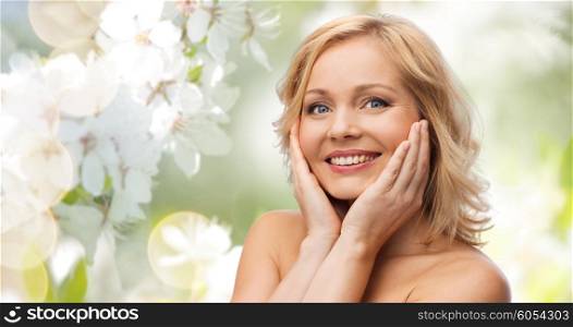 beauty, people, skincare and natural cosmetics concept - smiling woman with bare shoulders touching face over cherry blossom background
