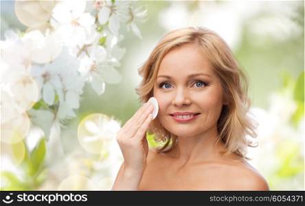 beauty, people, skincare and natural cosmetics concept - young woman cleaning face and removing make up with cotton pad over cherry blossom background