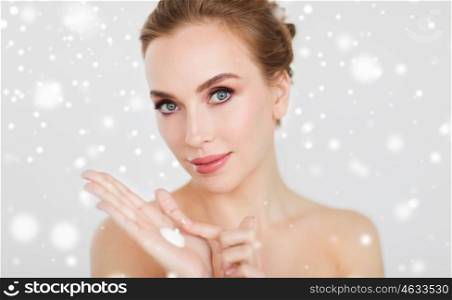 beauty, people, skincare, winter and cosmetics concept - close up of happy young woman with moisturizing cream on hand over gray background and snow