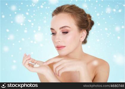 beauty, people, skincare, winter and cosmetics concept - happy young woman with moisturizing cream on hand over blue background and snow