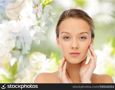 beauty, people, summer, organic and health concept - young woman with bare shoulders touching her face over green natural background with cherry blossom