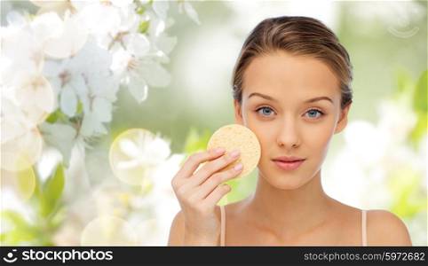 beauty, people, toiletry and skincare concept - young woman cleaning face with exfoliating sponge over cherry blossom background
