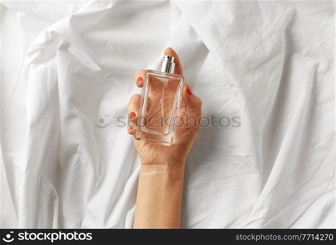 beauty, perfumery and people concept - female hand holding bottle of perfume on white sheet with folds. female hand with bottle of perfume on white sheet