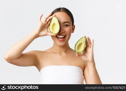 Beauty, personal care, spa and skincare concept. Beautiful happy asian woman in bath towel showing avocado pieces and smiling, nourish skin with face masks or daily cream, white background.. Beauty, personal care, spa and skincare concept. Beautiful happy asian woman in bath towel showing avocado pieces and smiling, nourish skin with face masks or daily cream, white background