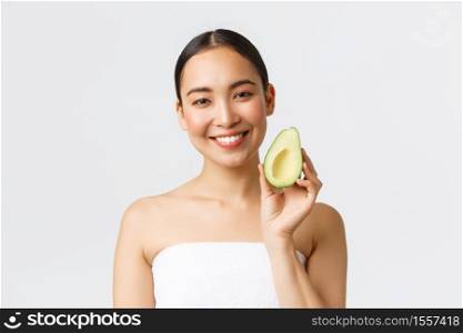 Beauty, personal care, spa and skincare concept. Close-up of attractive asian girl with fresh look standing in bath towel, showing avocado and smiling, recommend facial masks, moisturizing cream.. Beauty, personal care, spa and skincare concept. Close-up of attractive asian girl with fresh look standing in bath towel, showing avocado and smiling, recommend facial masks, moisturizing cream