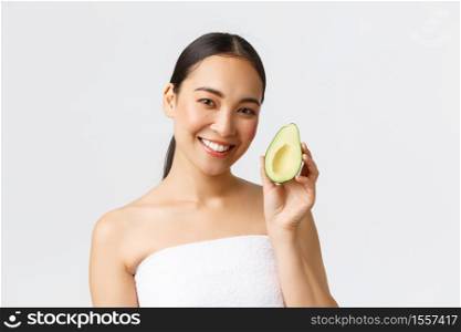 Beauty, personal care, spa and skincare concept. Close-up of beautiful asian female in bath towel showing avocado and smiling, recommend face mask or facial hydrating and nourishing cream.. Beauty, personal care, spa and skincare concept. Close-up of beautiful asian female in bath towel showing avocado and smiling, recommend face mask or facial hydrating and nourishing cream